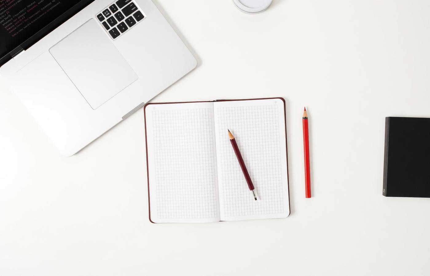 Overhead view of a laptop showing a terminal, an open grid notebook and a red pencil on a pale desk